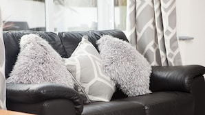 A black leather sofa with three grey pillows, a wooden coffee table, and a window with curtains. - The Nook (St Ives)