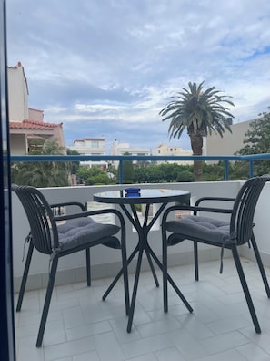 A balcony with a round table and two chairs, a palm tree, and buildings in the background. - Blue Roses (Rhodes)