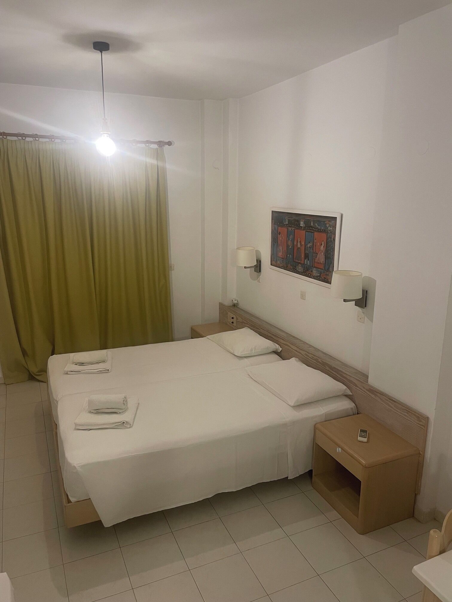 Hotelkamer met een bed, twee nachtkastjes, een wandlamp, een plafondlamp, een gordijn en een ingelijst kunstwerk aan de muur.