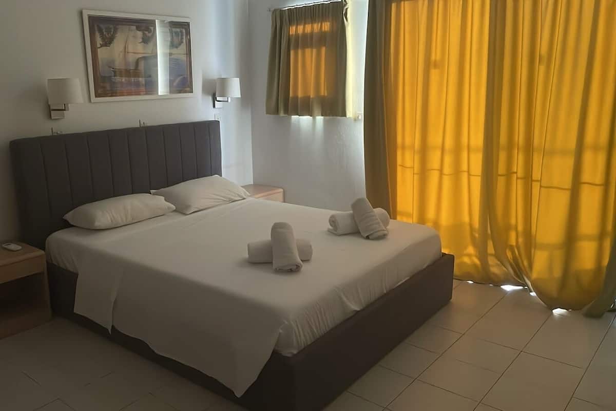 Una cama bien hecha con almohadas y toallas, una mesita de noche, una lámpara de pared y una ventana con cortinas.