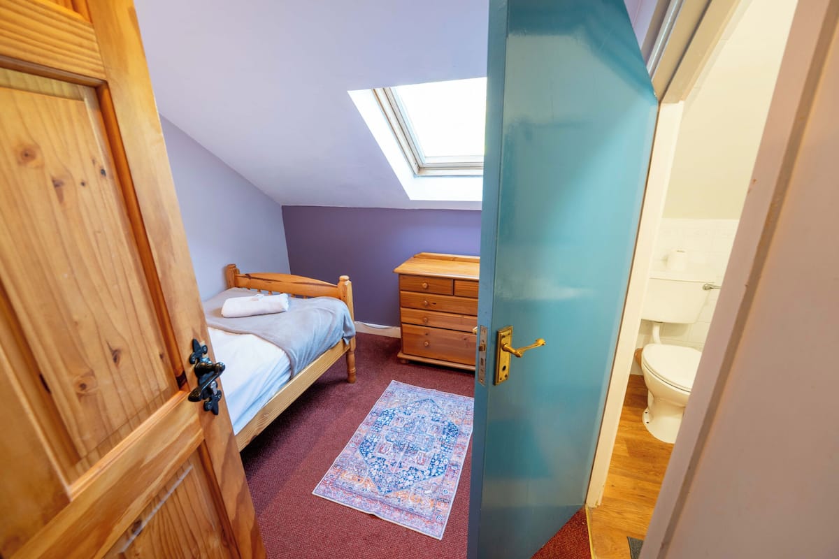 Une chambre avec un puits de lumière, un lit, une commode et une salle de bain visible à travers une porte ouverte.