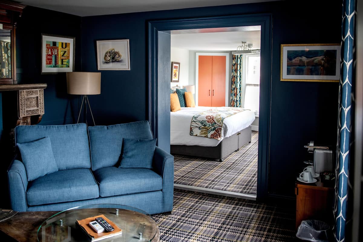 A hotel room with a blue sofa, a bed with a floral bedspread, a patterned floor, and a view into another room with a wardrobe and curtains.