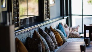 A cozy seating area with a bench and cushions, a table with a glass, and a window with blinds. - Marquis Of Granby (Dover)