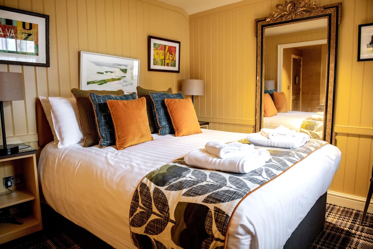 A neatly made bed with a patterned bedspread, multiple pillows, and a folded towel on the side. There is a bedside table with a lamp and a small electronic device. The room has a large mirror on the wall and framed artwork above the bed.