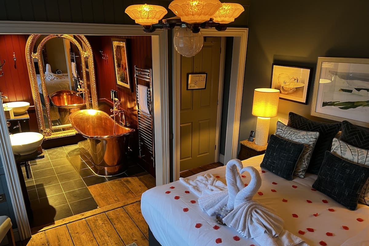 A hotel room with a large bed, a bathtub, a wooden floor, and a mirror with a gold frame.