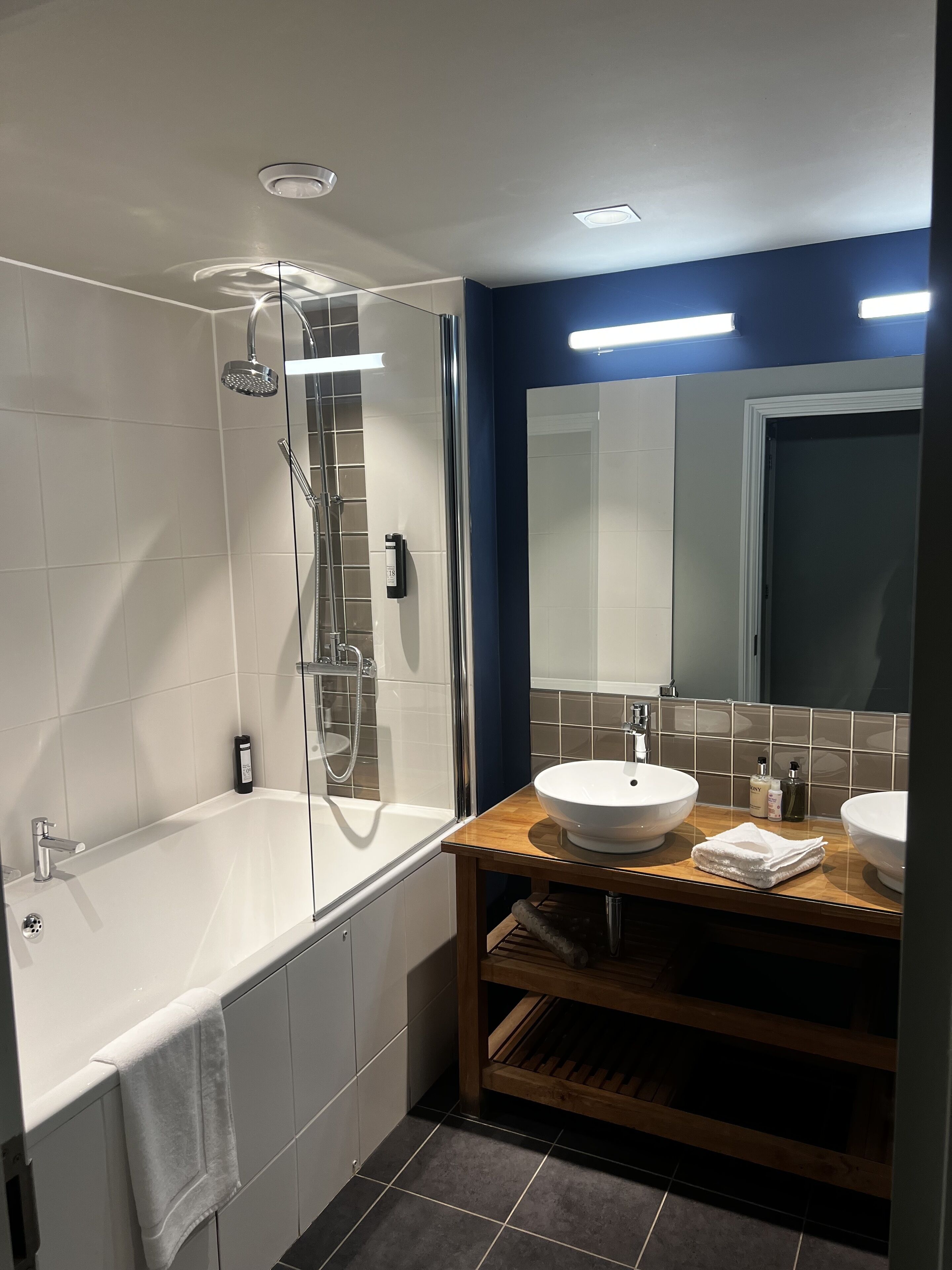 A modern bathroom with a glass-enclosed shower, a white bathtub, and two sinks with a wooden vanity.