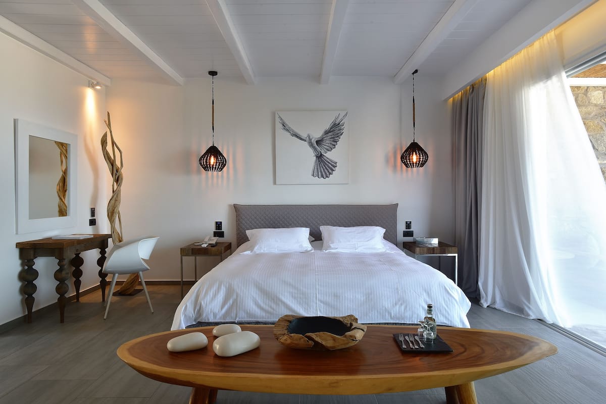 A modern hotel room with a large bed, a wooden table with a bowl and utensils, a chair, a desk with a lamp, and artwork on the walls.