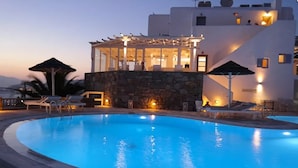 A swimming pool with lounge chairs and umbrellas, a stone wall, and a building with a covered outdoor area and lights.