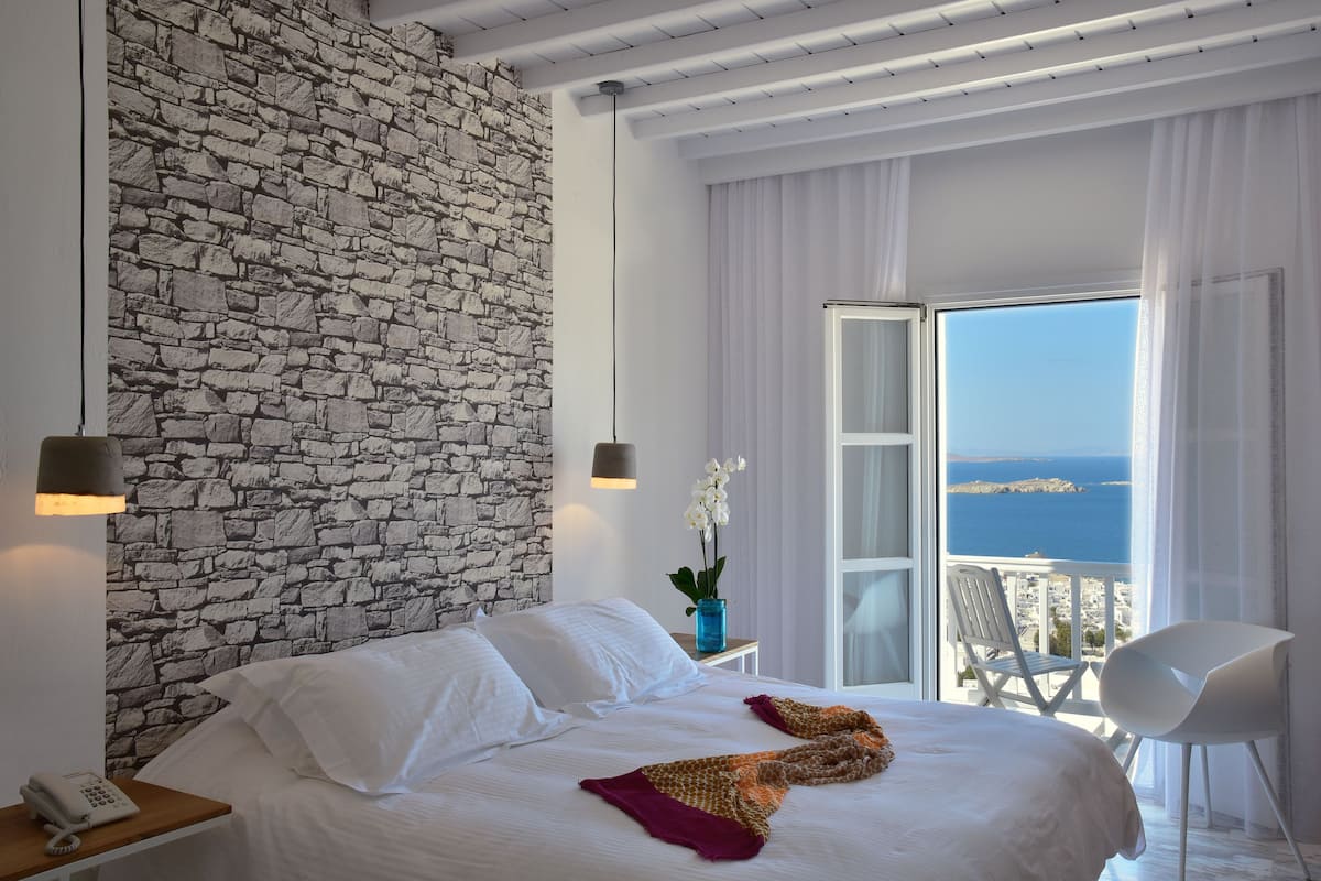 A bedroom with a stone wall, a bed with white linens, a nightstand with a phone, and a view of the sea through a balcony.