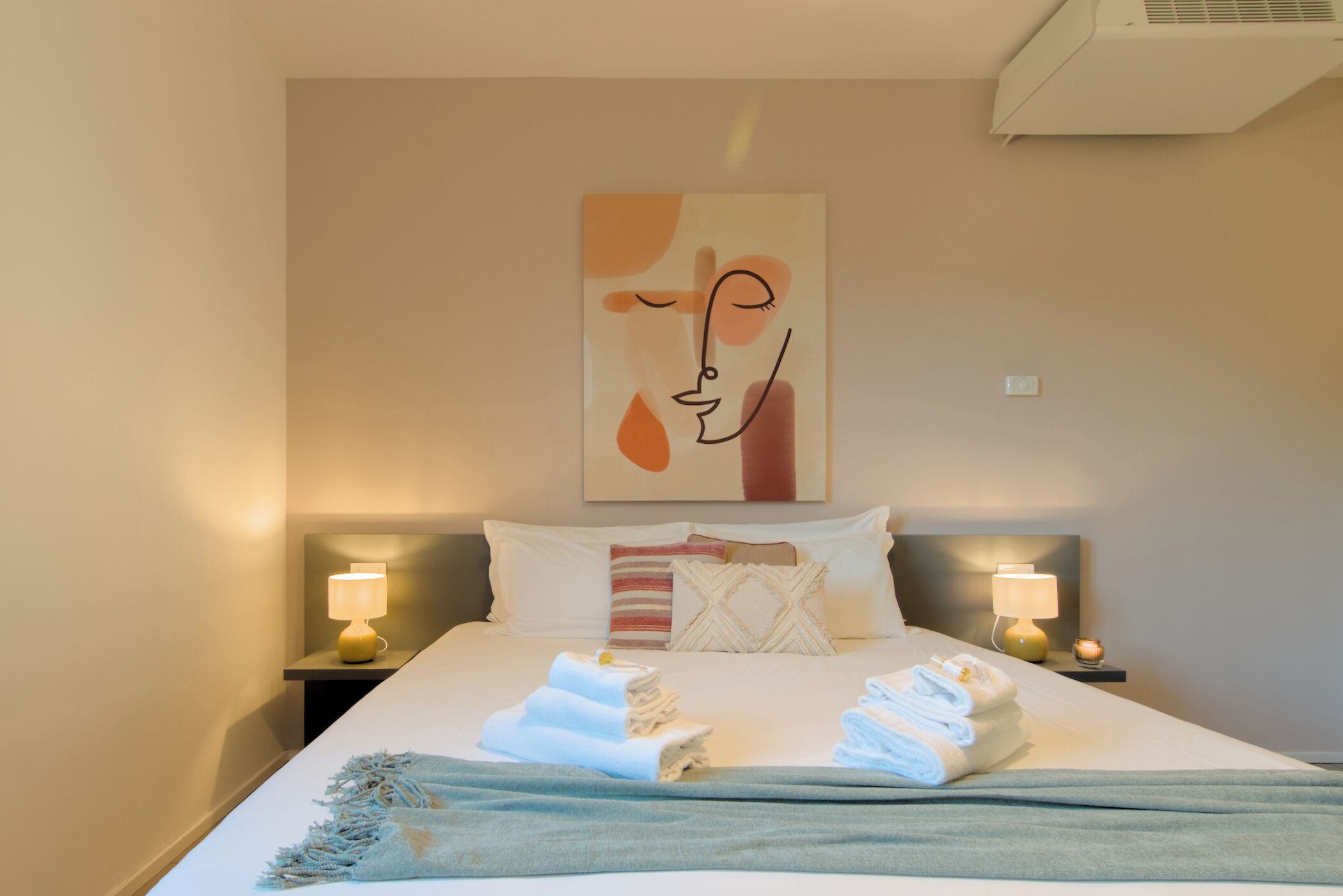 Een netjes opgemaakt bed met handdoeken, een kunstwerk aan de muur en twee bedlampjes.