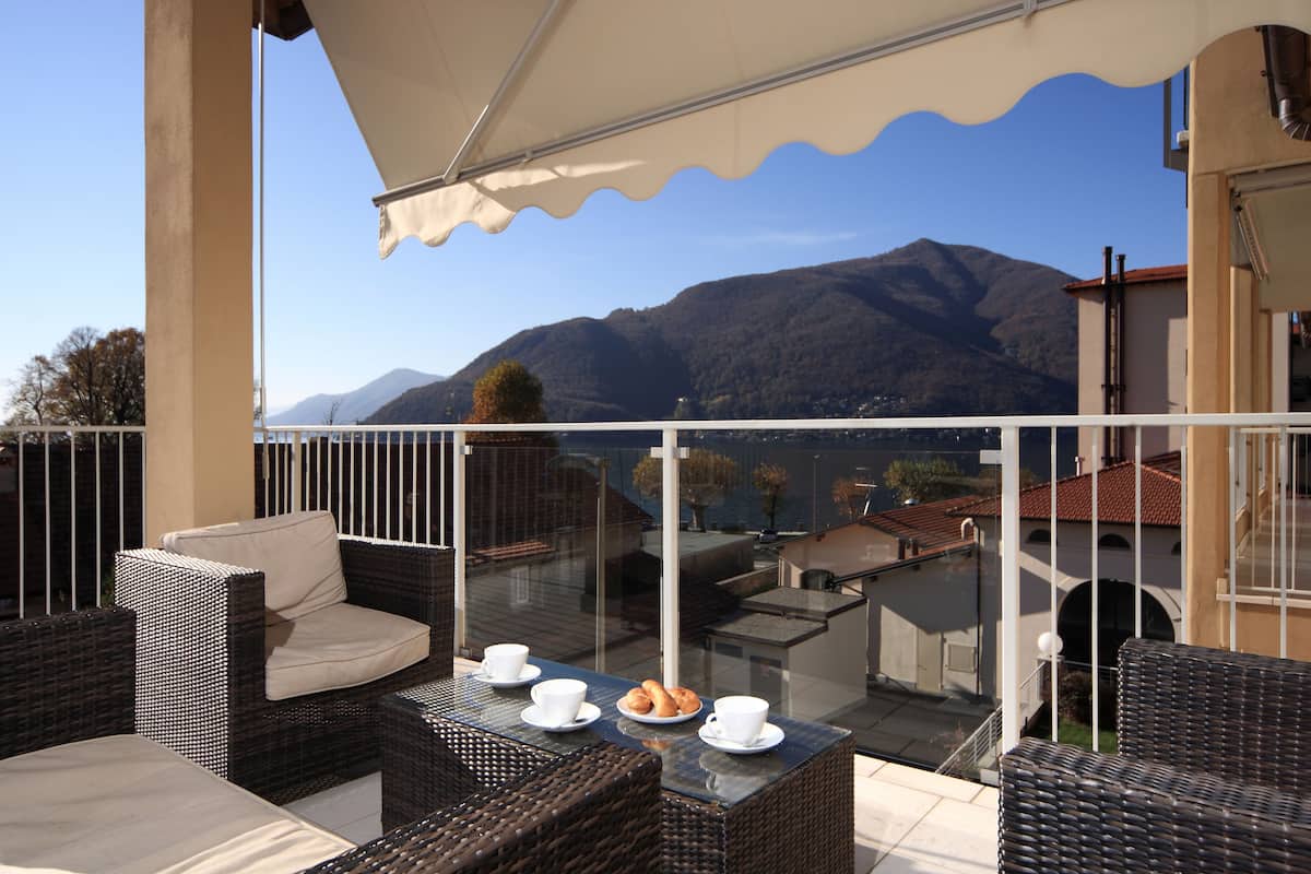 A balcony with wicker furniture, a glass table, and a view of mountains and a lake.