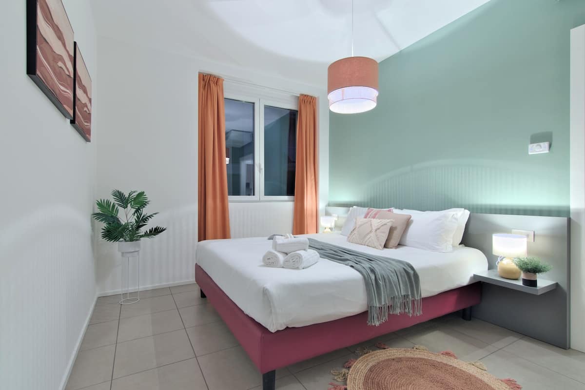 A bedroom with a bed, a bedside table with a lamp, a window with curtains, and a plant.