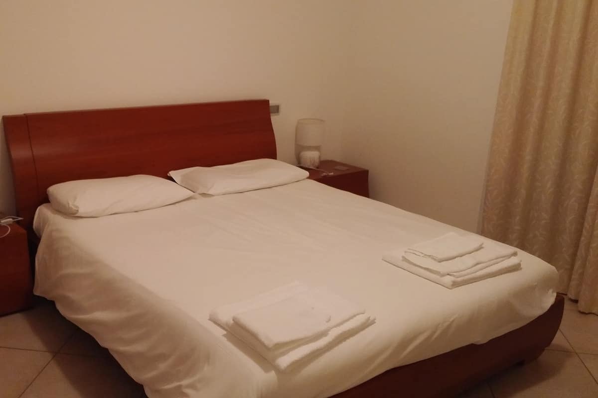 A neatly made bed with white linens and two pillows, a wooden headboard, a bedside table with a lamp, and a nightstand with a power outlet.
