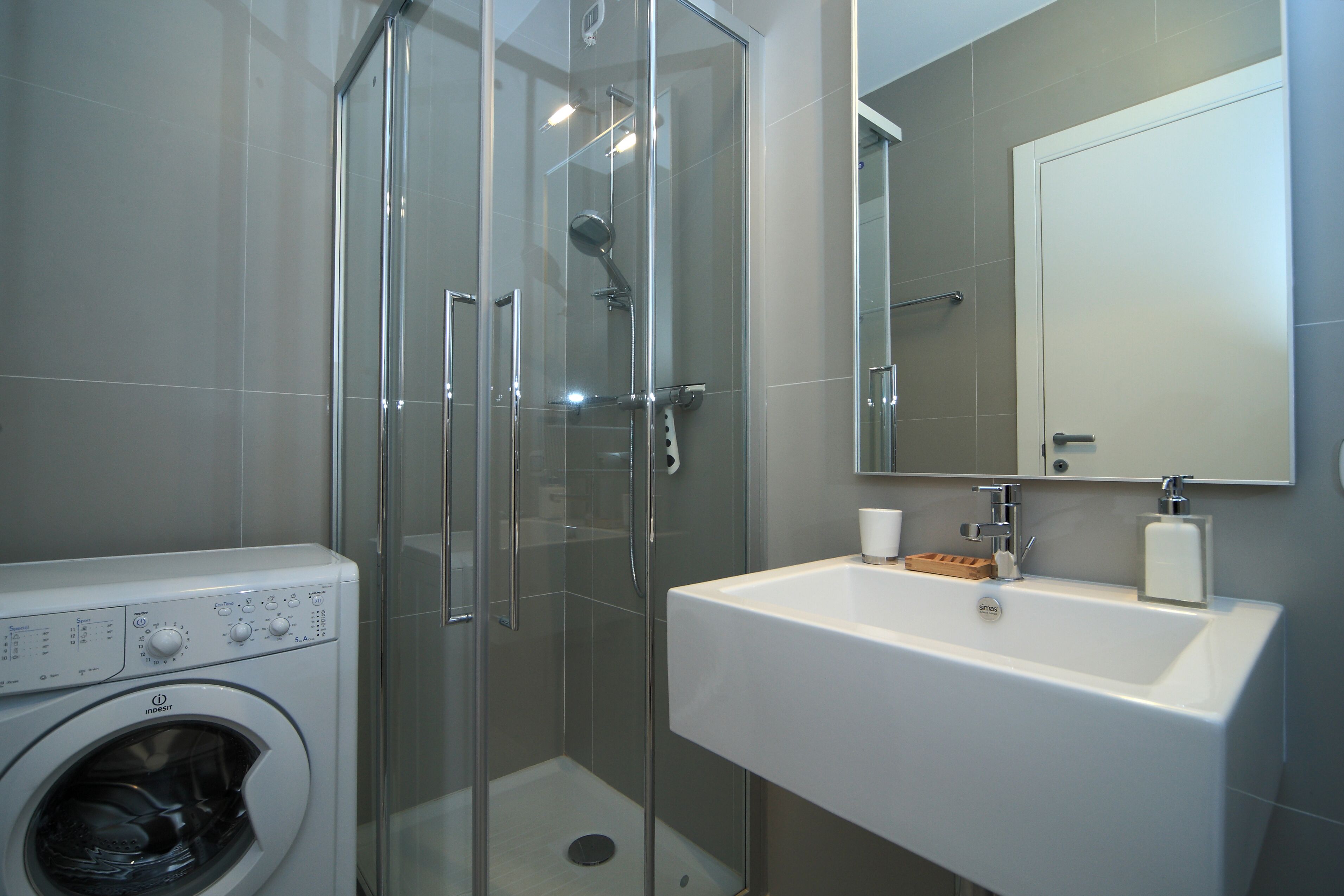 A modern bathroom with a glass-enclosed shower, a built-in washing machine, and a white sink with a mirror above it.