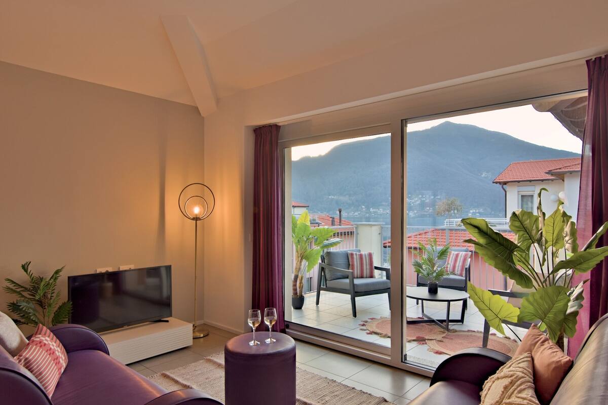 A modern living room with a flat-screen TV, comfortable seating, and a large sliding glass door leading to a balcony with a mountain view.