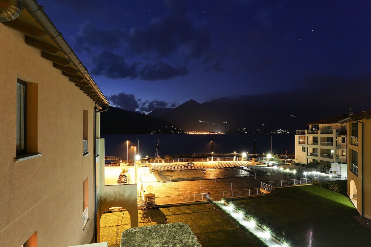 A nighttime view of a coastal area with a parking lot, a building with a sloped roof, and a distant mountain range.