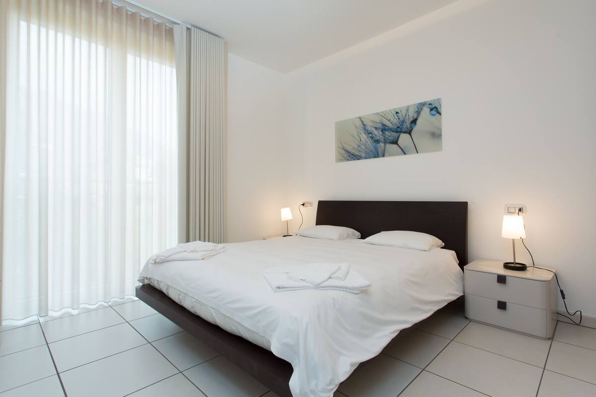 A modern hotel room with a large bed, two bedside tables with lamps, a wall-mounted artwork, and a large window with sheer curtains.