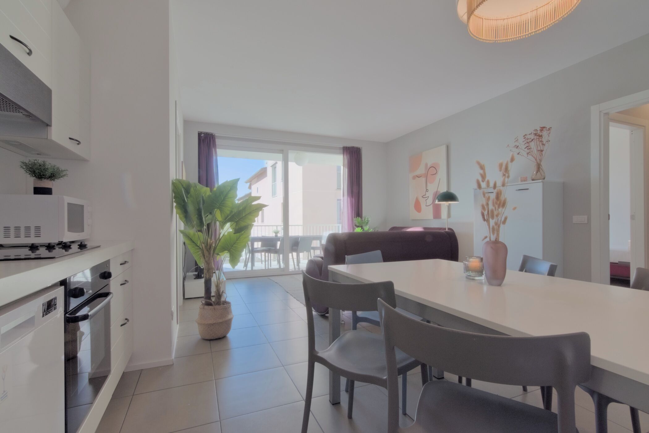 Een moderne keuken met witte kastjes, een magnetron en een oven. Er is een eethoek met een witte tafel en stoelen, en een grote potplant.