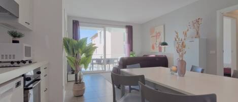 Een moderne keuken met witte kastjes, een magnetron en een oven. Er is een eethoek met een witte tafel en stoelen, en een grote potplant.