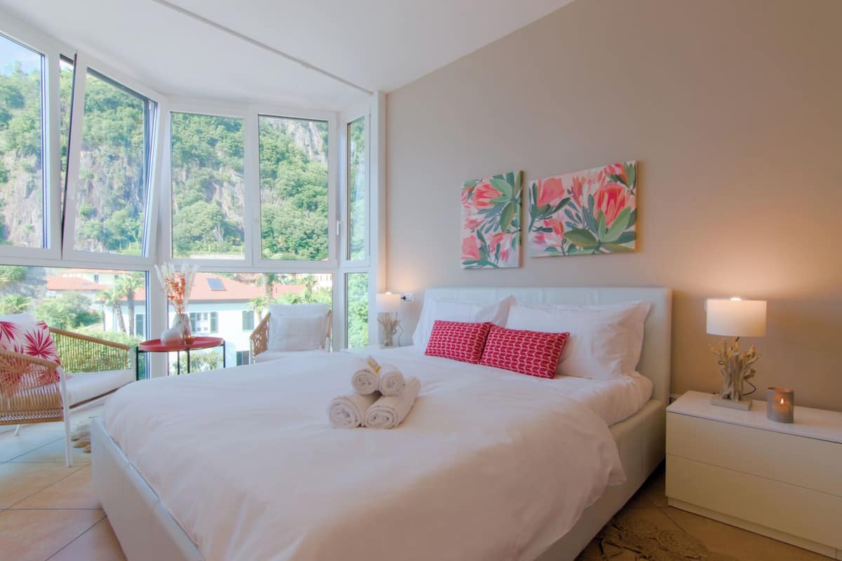 A spacious bedroom with a large bed, two bedside tables, and a view of greenery through large windows.