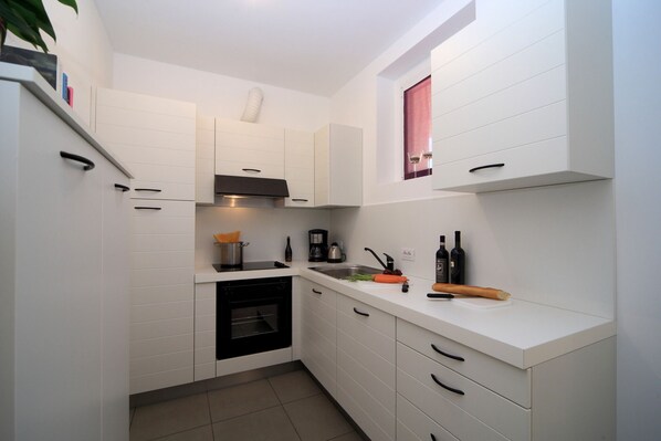 A compact kitchen with white cabinetry, a built-in oven, and a countertop with a coffee maker, wine bottles, and a rolling pin.