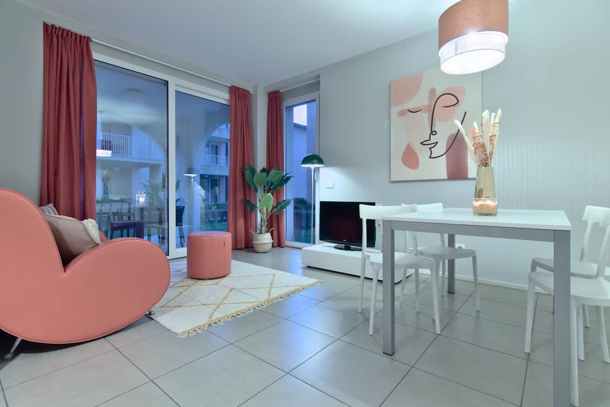 A modern living room with a pink sofa, a dining table with white chairs, a flat-screen TV, a potted plant, and a large window with curtains.