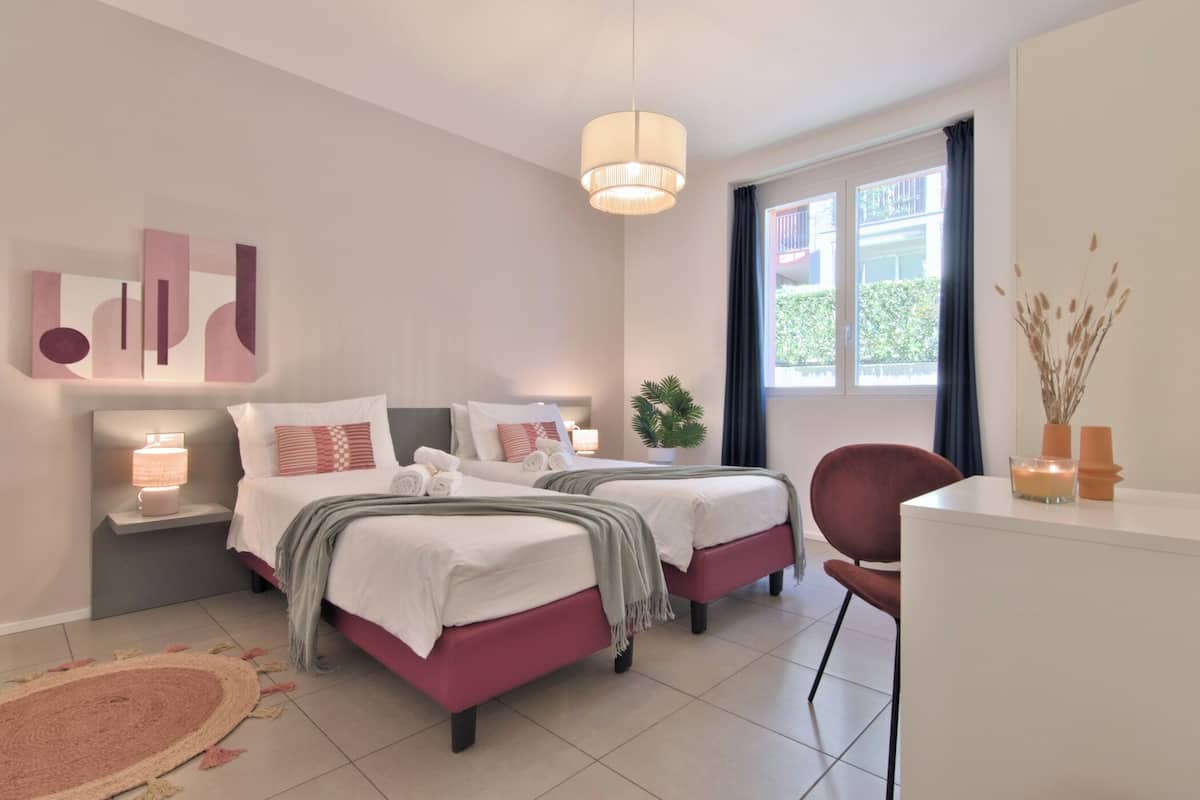 A modern bedroom with two beds, a bedside table with a lamp, a chair, and a window with curtains.
