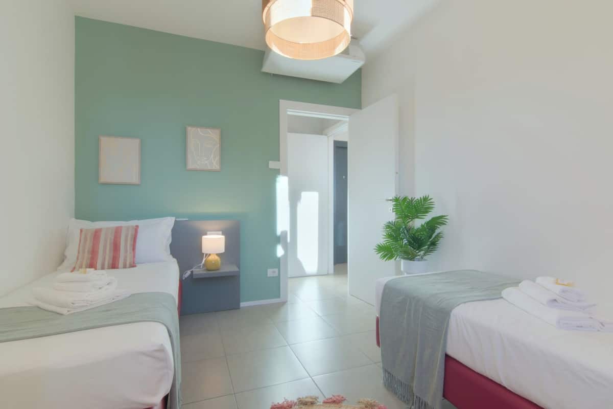A hotel room with two beds, a small table with a lamp, a potted plant, and a view of the hallway.