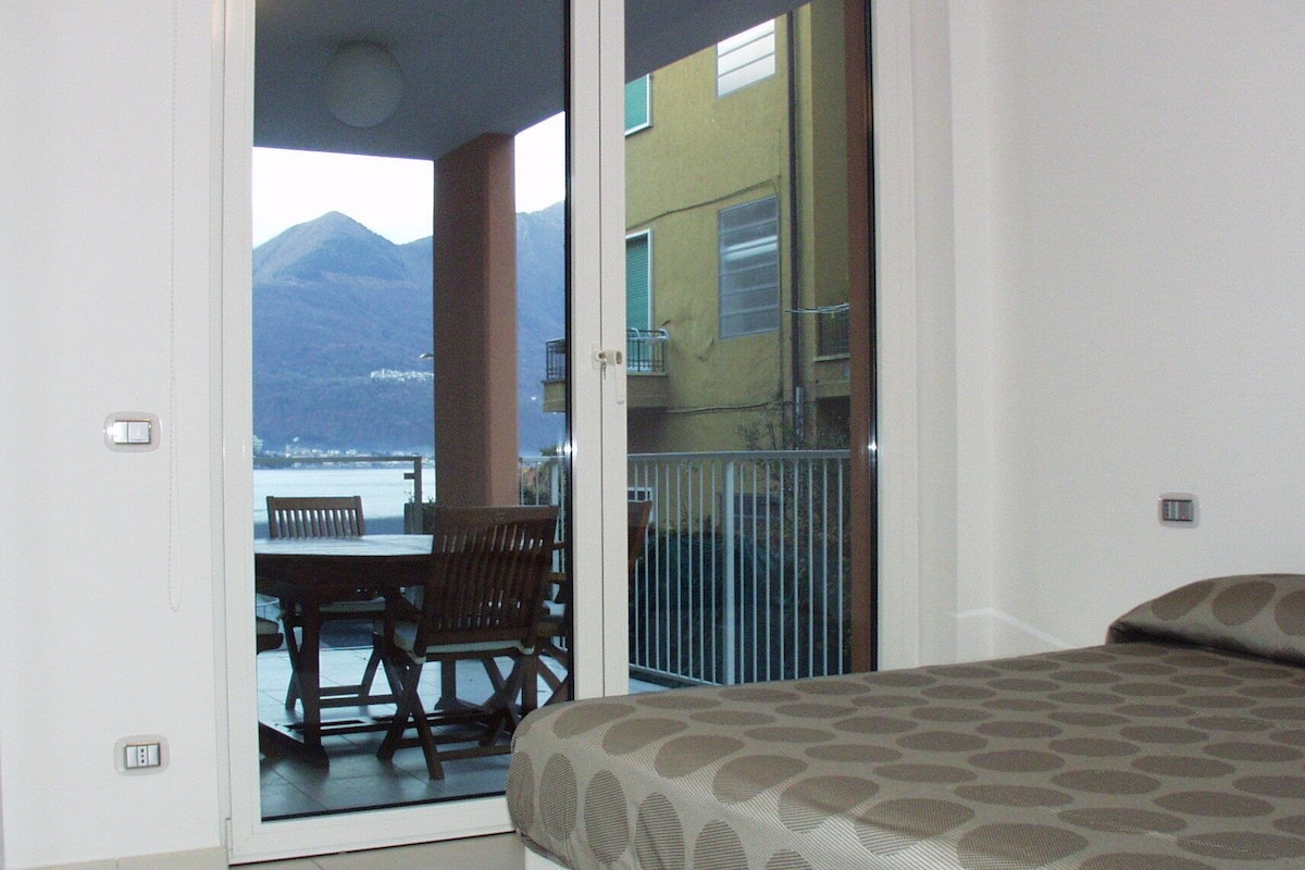 A bedroom with a bed, a balcony with a table and chairs, and a view of a mountain and water.