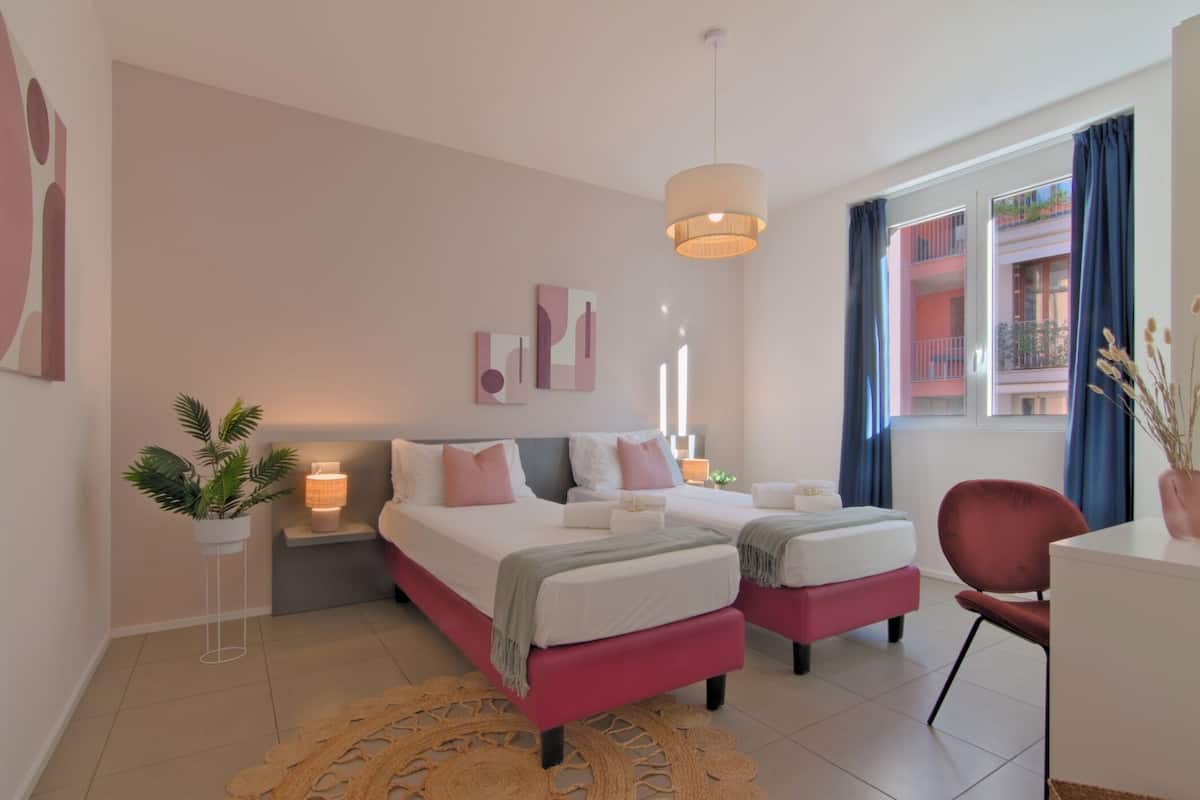 A modern bedroom with two beds, a desk, and a chair. There are wall art pieces and a potted plant.
