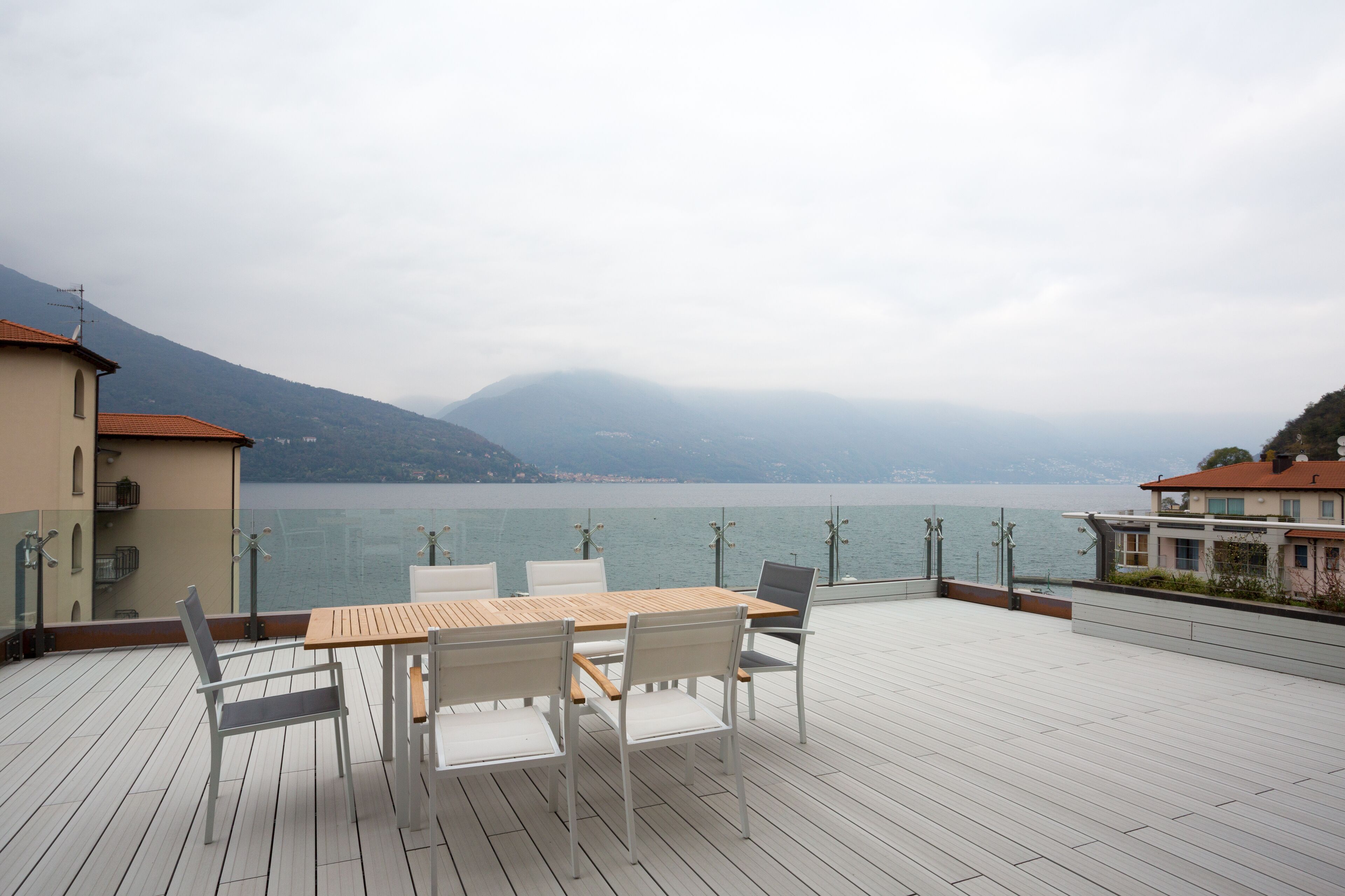 A wooden deck with a table and chairs, overlooking a lake with mountains in the background.