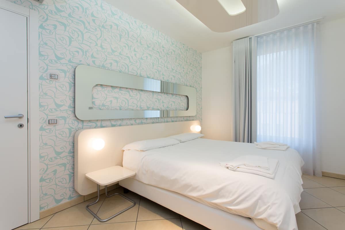 A modern hotel room with a large bed, a bedside table, a built-in bench, a mirror, and a window with curtains.
