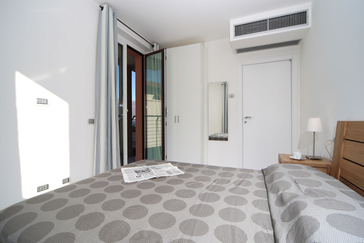 A bedroom with a bed, a nightstand, a lamp, a wardrobe, a door, and an air conditioning unit.