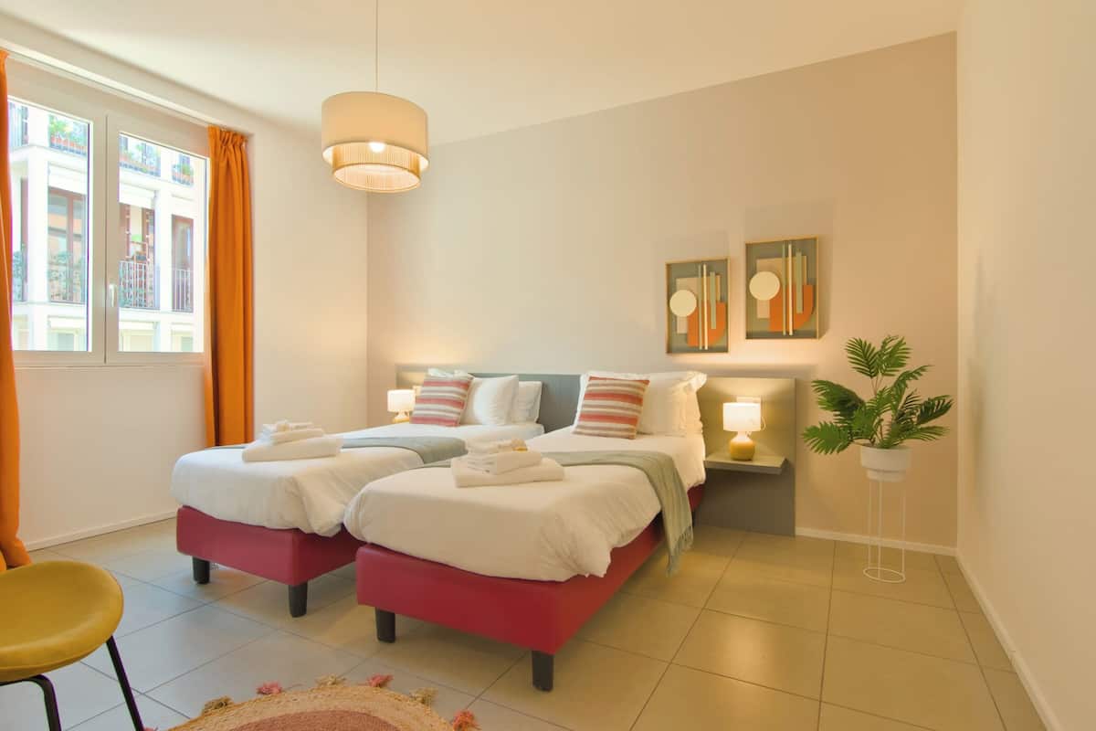A double bed with a red base and white bedding, two bedside tables with lamps, a potted plant, and a view of buildings through a window with orange curtains.