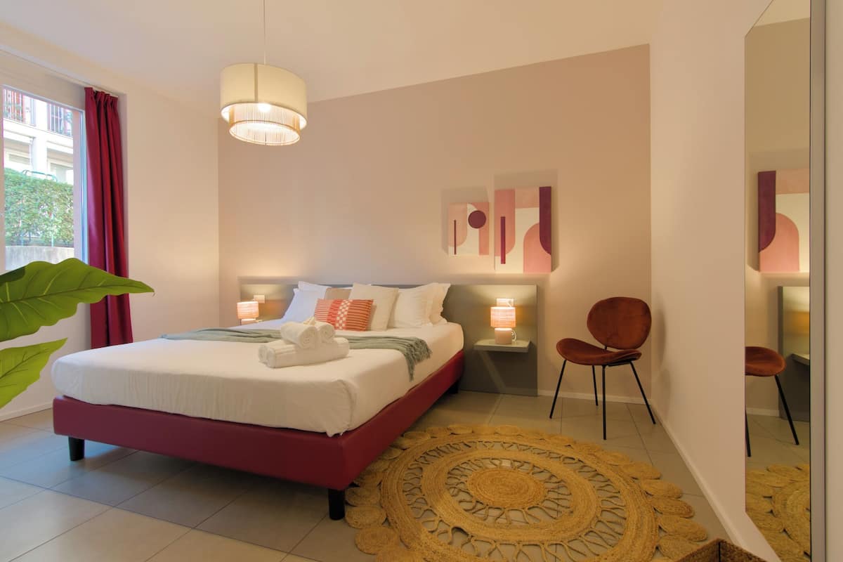 A modern bedroom with a bed, bedside lamps, a large window with curtains, a round woven rug, and a chair.