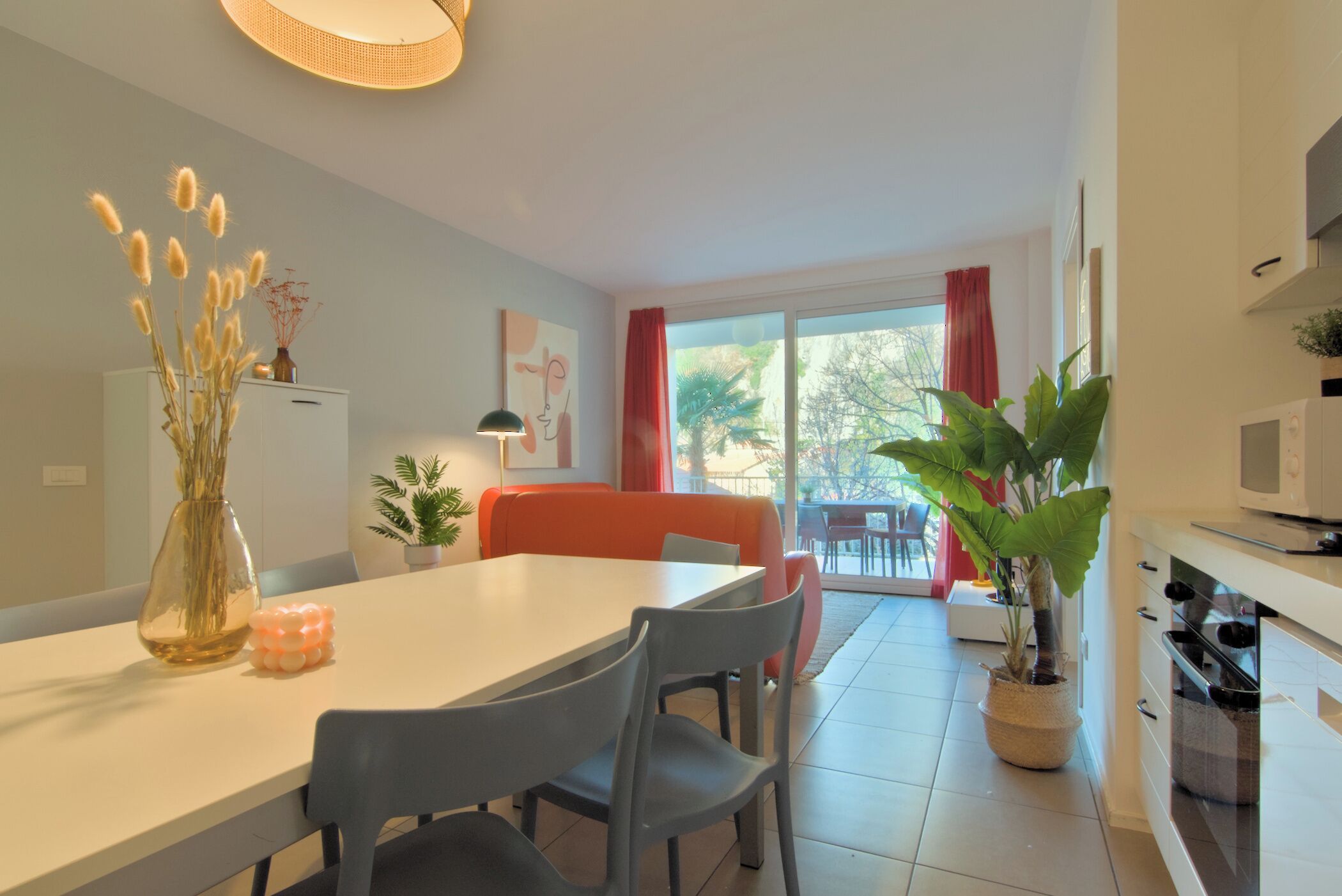 A modern kitchen with white cabinets, a microwave, and a stove. There is a dining table with chairs and a vase with dried plants. A large window with red curtains is visible in the background.