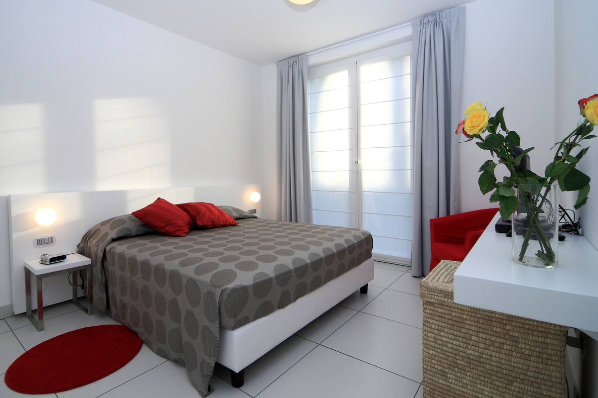 A modern bedroom with a bed, bedside tables, a red rug, a red chair, and a vase with flowers.