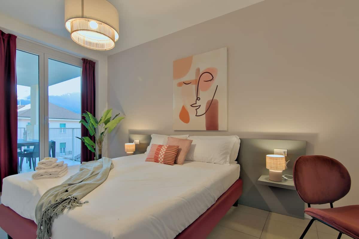 A modern hotel room with a large bed, a bedside table with a lamp, a chair, a potted plant, and a view of the outdoors.