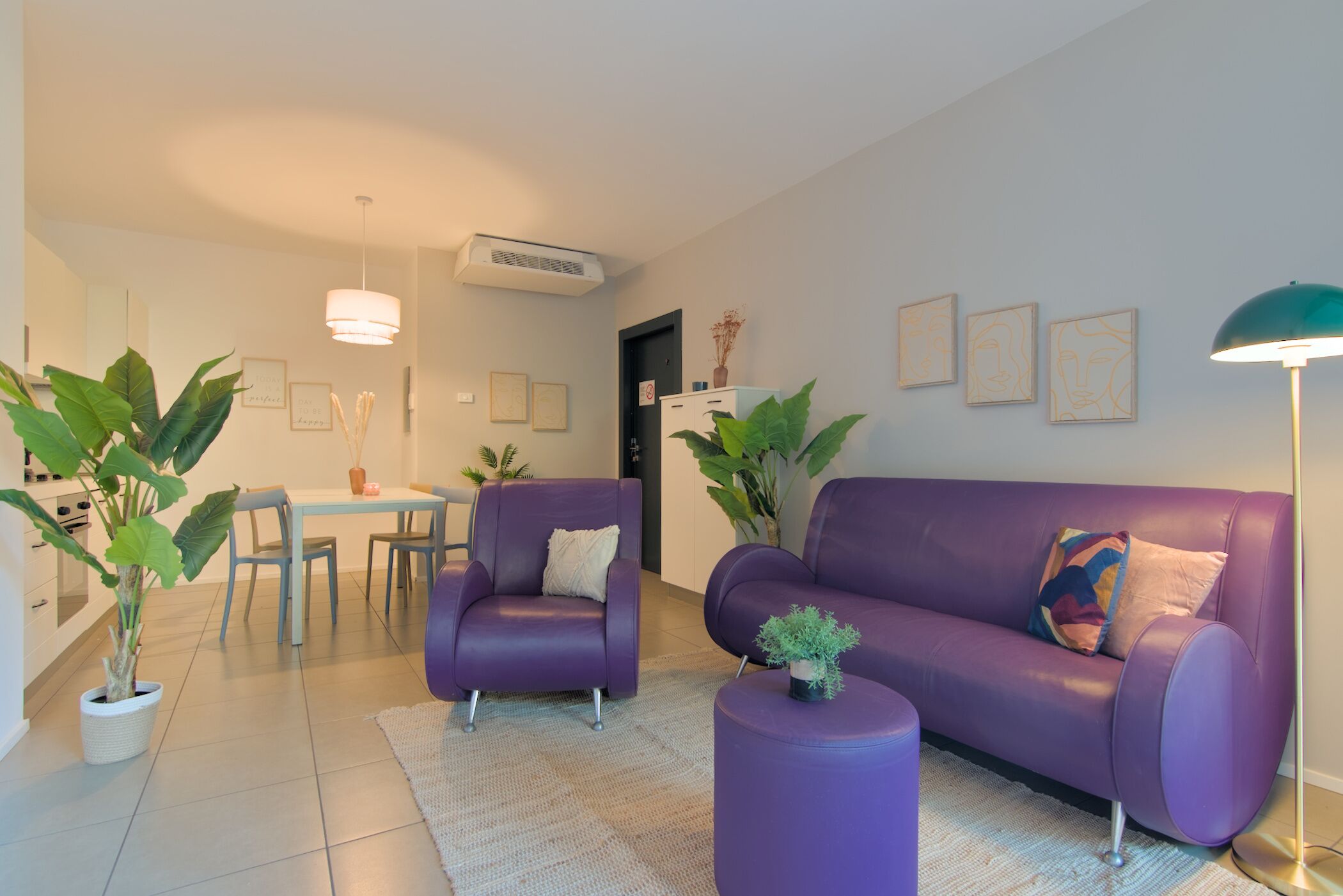 A modern living room with a purple sofa, matching armchair, and a small table with a plant. There is a dining area with a white table and chairs, and a kitchen area with a refrigerator and a potted plant.