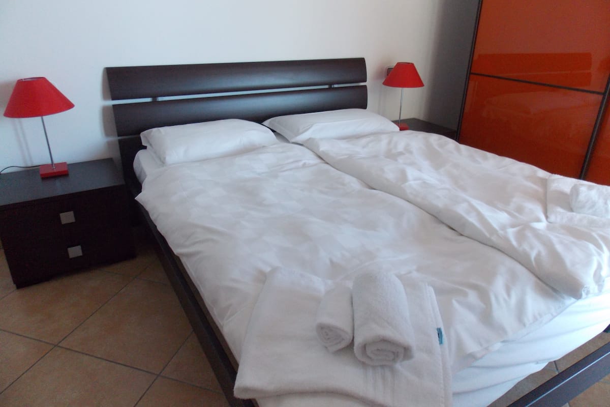 A neatly made bed with white linens, two red lamps on nightstands, and a red wall panel.