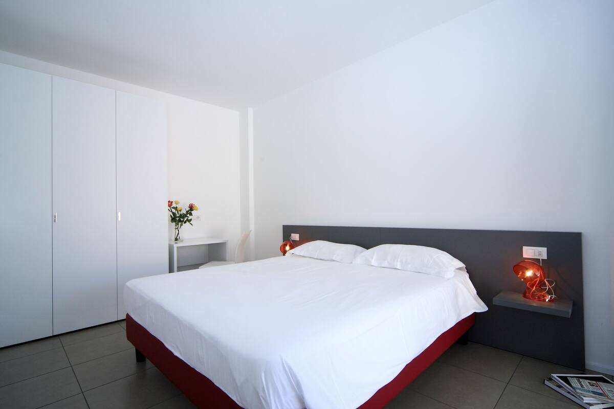 A modern bedroom with a large bed, white walls, and a red accent on the bed frame.