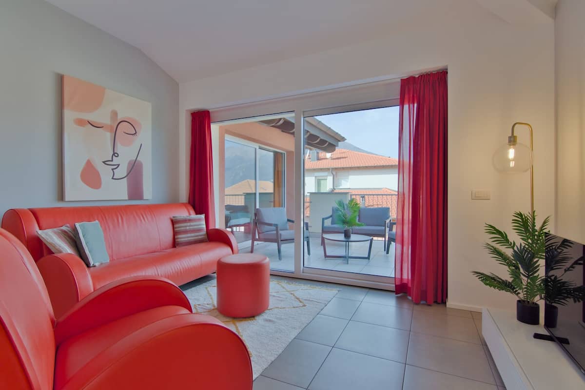 A modern living room with a red sofa, a coffee table, and a TV on a stand. There is a sliding glass door leading to an outdoor patio area with a table and chairs.