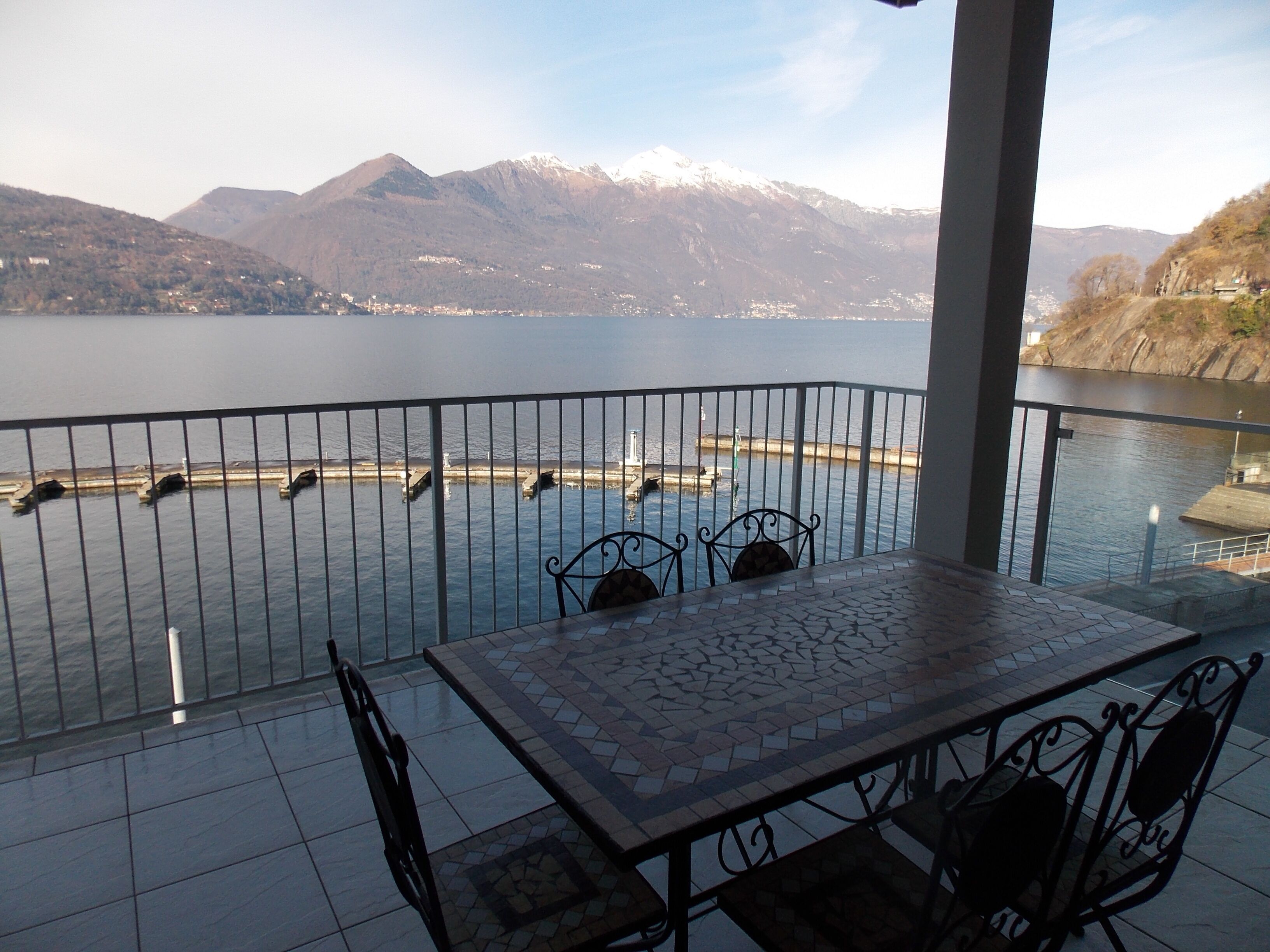 A balcony with a view of a lake and mountains. There is a table with chairs and a railing.