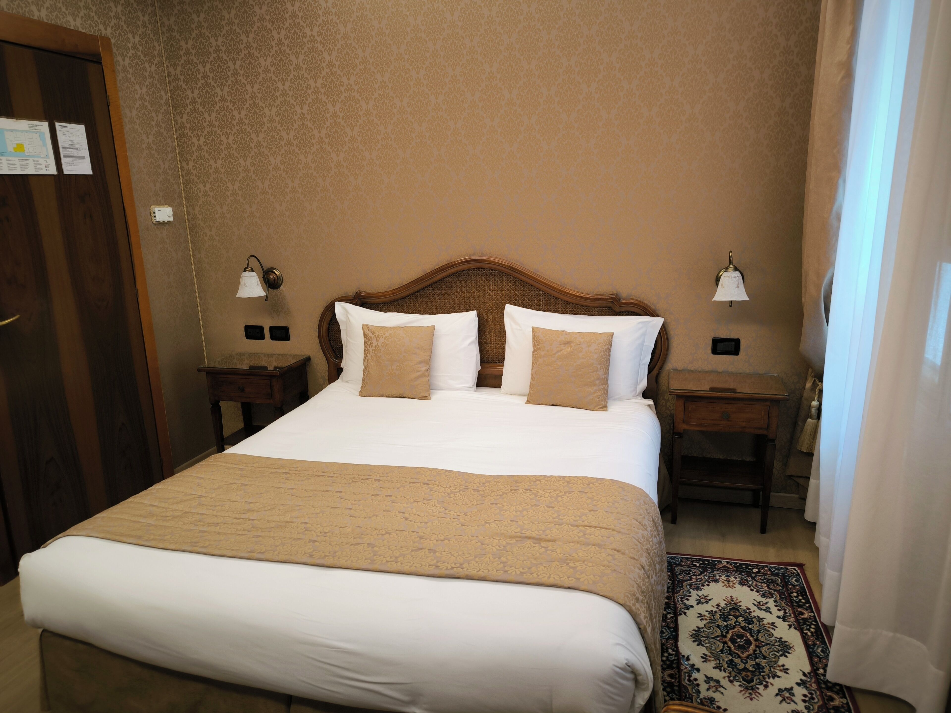 Een opgemaakt bed met een beige sprei, twee nachtkastjes met lampen en een kamerbreed vloerkleed.