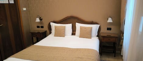 Een opgemaakt bed met een beige sprei, twee nachtkastjes met lampen en een kamerbreed vloerkleed.