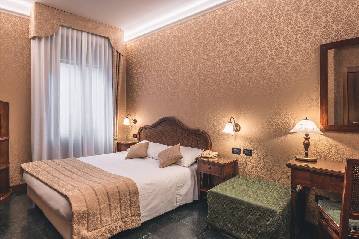 Habitación de hotel con una cama grande, dos mesitas de noche con lámparas, un escritorio pequeño con una silla y una ventana con cortinas.