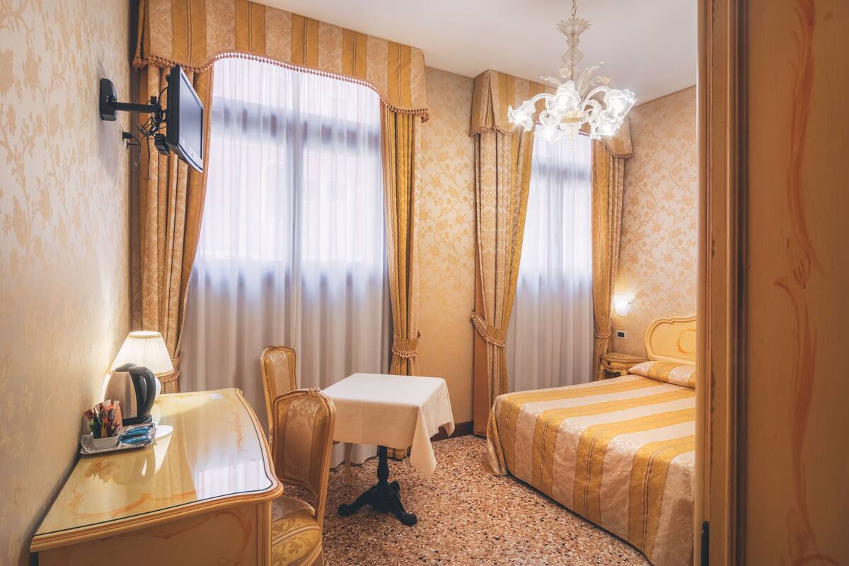 Habitación de hotel con cama, una mesita, una silla, escritorio, lámpara y candelabro.