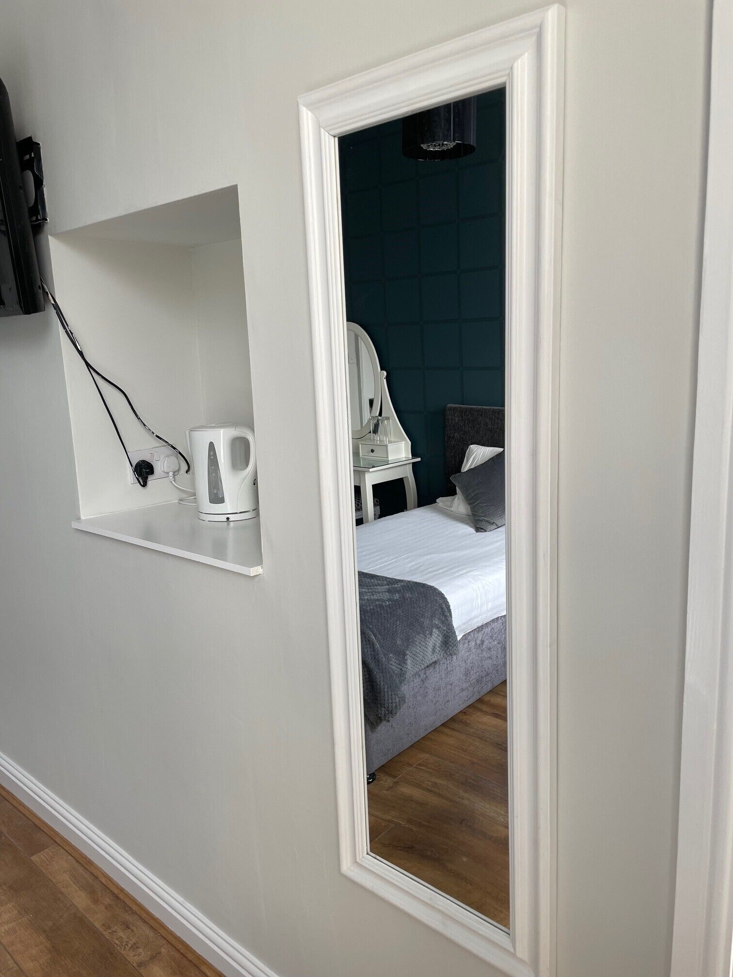 Une chambre à coucher comprenant un lit, une table de chevet, un miroir et une étagère avec une bouilloire.