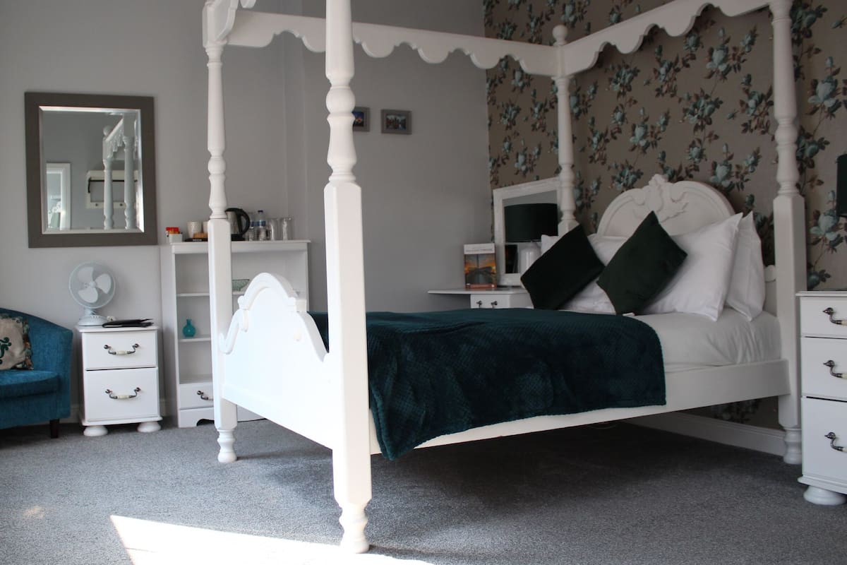 A bedroom with a four-poster bed, a bedside table, a chair, a dresser, and a mirror.