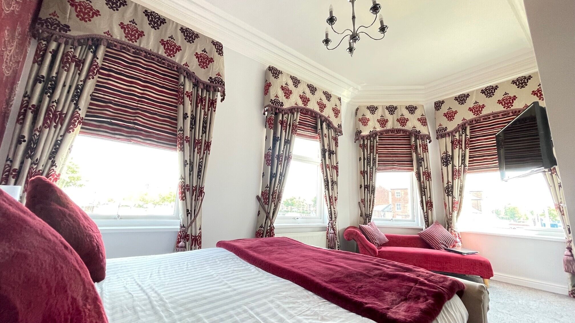 Un dormitorio con una cama grande, dos camas individuales y un chaise lounge. Hay varias ventanas con cortinas, un candelabro y vistas al exterior.